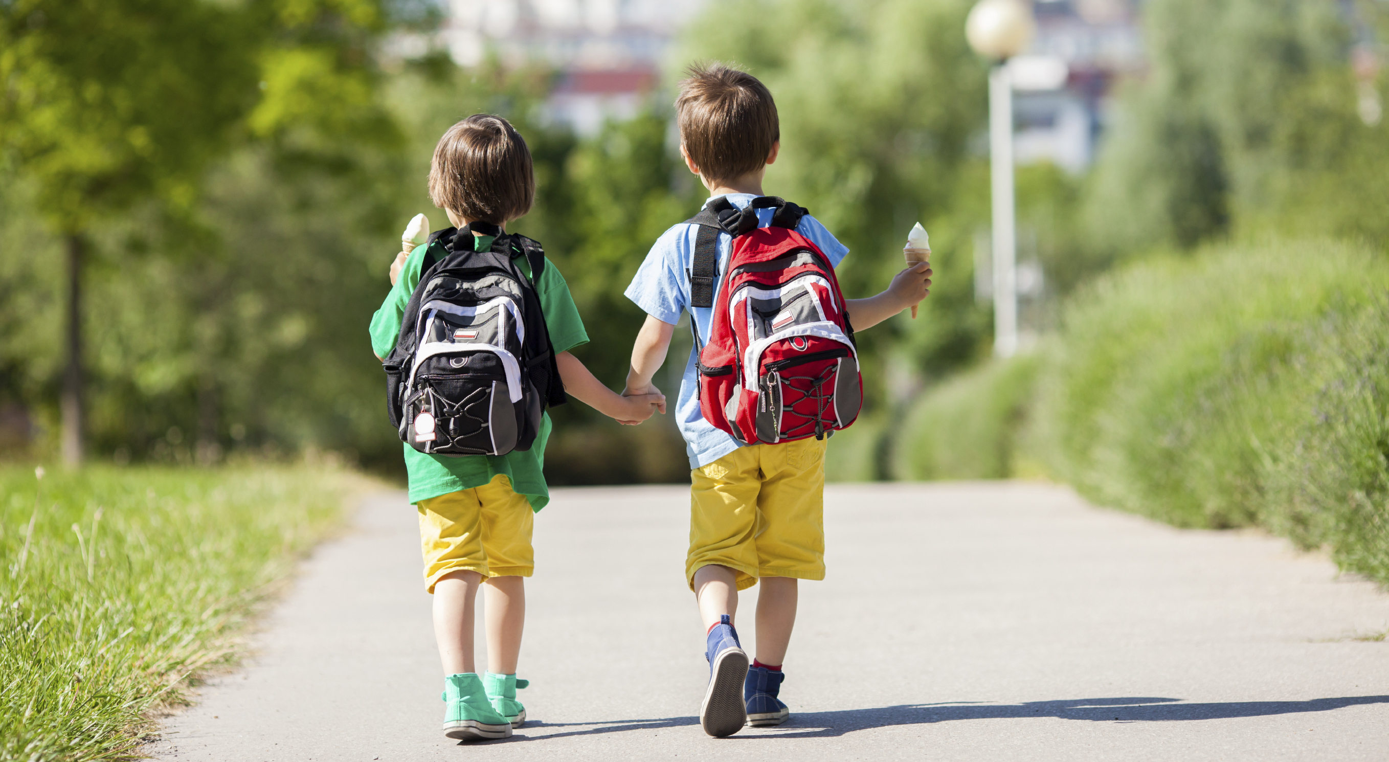 Two adorable boys in colorful clothes and backpacks, walking away, holding and eating ice cream on a sunny summer afternoon, warm day, casual clothing