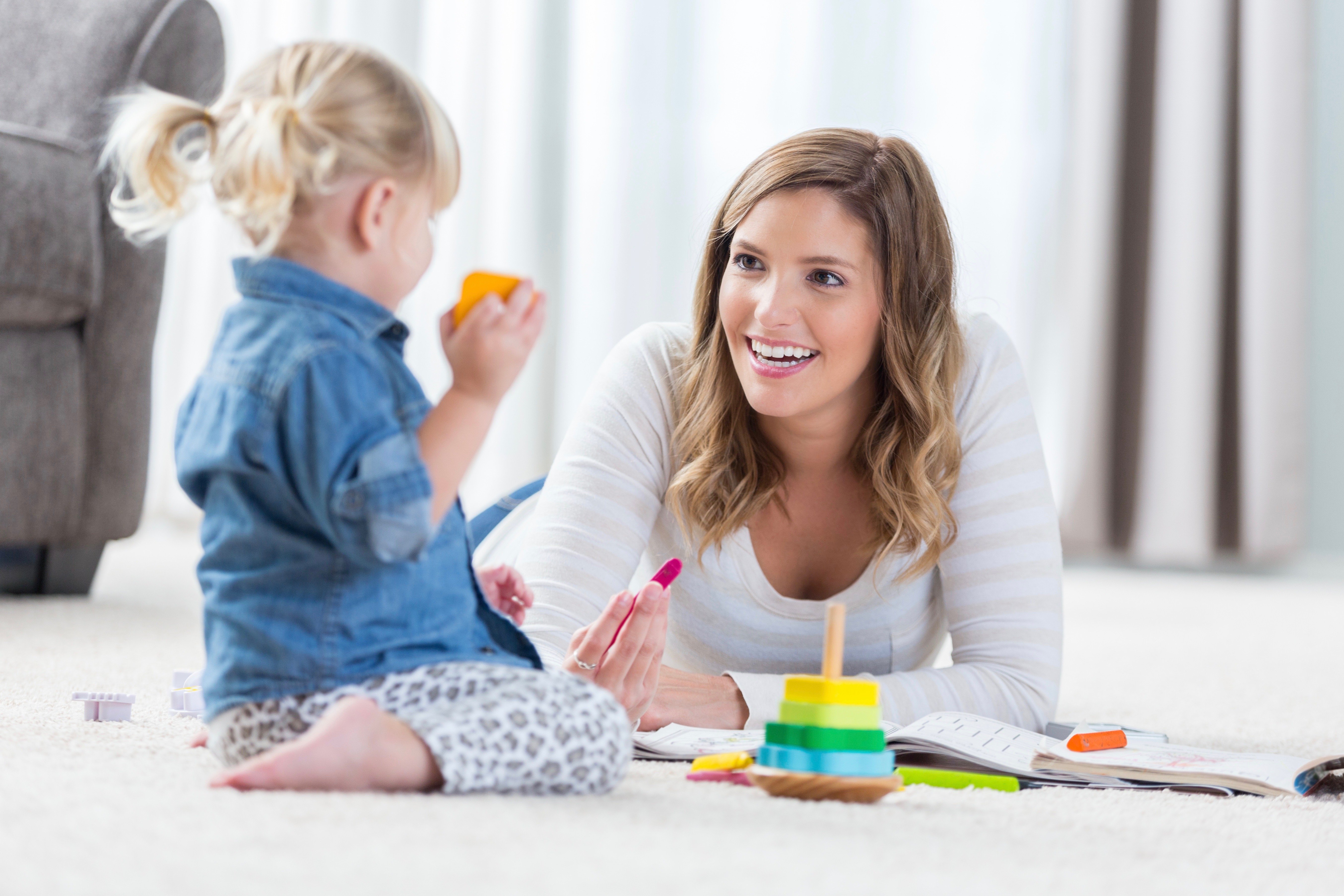 Happy babysitter and preschool age girl play with toys and color in a coloring book together. They are playing on the floor in their home.